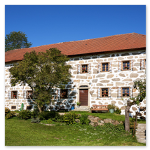 Großes Steinbloßhaus mit roten Ziegeldach, Fensterläden, Vorgarten und Holzbank; sonnige ländliche Szene in Stiftung bei Neumarkt im Mühlviertel