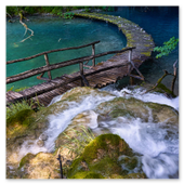 Blick von oben auf einen kleinen Wasserfall, der direkt unter einem geschwungenen Holzsteg hindurchfließt, welcher über das tiefblaue und türkisfarbene Wasser im Nationalpark Plitvicer Seen führt.