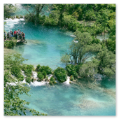 Vogelperspektive auf eine hölzerne Aussichtsplattform mit einer Touristengruppe an einem glasklaren, türkisfarbenen Flusslauf mit kleinen Wasserfällen und grünen Bäumen im Nationalpark Plitvicer Seen.