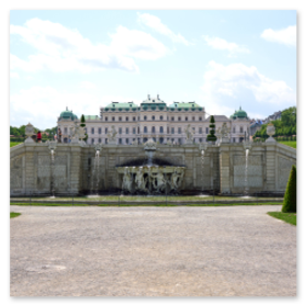 Das Foto zeigt den historischen Belvedere-Palast in Wien, Österreich. Der eindrucksvolle Bau mit mehreren grünen Kuppeln und kunstvollen Fassadendetails steht im Zentrum des Bildes. Davor erstreckt sich ein großer Brunnen mit Skulpturen und Wasserfontänen, umgeben von gepflegten Gartenanlagen und symmetrischen Wegen. Die Szene wirkt durch das klare Blau des Himmels besonders lebendig und strahlend. Es ist ein typisches Beispiel für barocke Architektur und Wiener Kulturerbe.