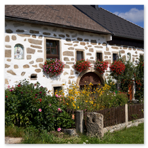 Traditionelles Steinbloßhaus mit dunklem Schindeldach, Rundbogentor, Fensterblumen und blühendem Vorgarten; ländliche Szene in Ottenschlag im Mühlviertel