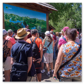 Eine große Gruppe von Touristen steht von hinten fotografiert vor einer großen, hölzernen Informationstafel mit einer geografischen Übersichtskarte des Plitvicer Seen Nationalparks an einem sonnigen Tag.