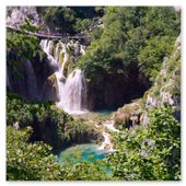 Blick durch grüne Blätter auf einen großen, mehrstufigen Wasserfall im Nationalpark Plitvicer Seen, der in ein türkisfarbenes Becken stürzt, mit einem hölzernen Wanderweg voller Besucher im Hintergrund.