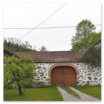 Drei verbundene Steinbloßgebäude mit roten Ziegeldächern, dunklen Fensterläden und großem Rundbogentor; Hochbeete, Kiesweg und ländliche Umgebung in Stiftung bei Neumarkt im Mühlviertel
