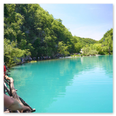 Touristen wandern auf einem hölzernen Steg entlang einer steilen Felswand direkt am Ufer eines intensiv türkisblauen, spiegelglatten Sees im Nationalpark Plitvicer Seen unter blauem Himmel.