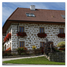 Traditionelles Steinbloßhaus mit roten Ziegeldach, Fensterblumen und gepflegtem Garten; gepflasterter Zugang und ländliche Umgebung in Ottenschlag im Mühlviertel
