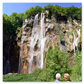 Zwei Personen betrachten von unten den beeindruckenden Veliki Slap Wasserfall im Nationalpark Plitvicer Seen, dessen Wasser erhaben über eine hohe, bewachsene Felswand in die Tiefe stürzt.