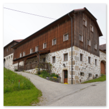 Großes Steinbloßhaus mit dunklem Holzaufbau, Rundbogentor und Balkon; geschwungene Straße, Wiesen mit Wildblumen und weitere Höfe in Stiftung bei Neumarkt im Mühlviertel