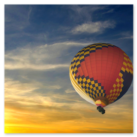 Heißluftballon mit rot-gelber-blauer Karomuster-Hülle schwebt bei Sonnenaufgang über einem wolkigen Himmel. Der warme Sonnenaufgangshorizont taucht die unteren Wolkenschichten in orange-goldene Farbtöne, während die obere Atmosphäre in kühlen Blautönen verläuft. Ideal für SEO-Stichworte wie Heißluftballon, Sonnenaufgang, Morgenstimmung, Panorama, Luftfahrt, Abenteuer und Naturfotografie.