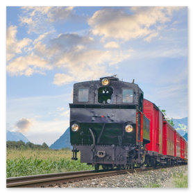 Szene eines Personenzugs mit schwarzer Lokomotive und roten Waggons, der auf einer eingleisigen Bahnstrecke entlang einer Parallelstraße durch eine grüne, hügelige Landschaft mit im Hintergrund sichtbaren Bergen und blauem, leicht bewölktem Himmel fährt. Ideal für SEO-Stichworte wie Regionalzug, Alpenbahn, Bahnstrecke, Landschaftsfotografie, Zugfahrt, Bergpanorama und Mobilität.