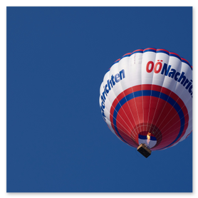 Fotografie eines Heißluftballons der OÖNachrichten mit weißer Hülle, roten und blauen Streifen sowie dem Schriftzug „OÖNachrichten“, über dem Korb sichtbar eine brennende Gasflamme, vor einem tiefblauen Himmel. Ideal für SEO-Stichworte wie Heißluftballon, OÖNachrichten, Ballonfahrt, Luftfahrtfotografie, Abenteuer und Sommererlebnis.