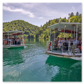Zwei mit Touristen besetzte Elektroboote fahren an einem sonnigen Tag über das ruhige, grüne Wasser des Kozjak-Sees im Nationalpark Plitvicer Seen, umgeben von bewaldeten Hügeln.