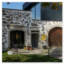 Gebäude mit traditioneller Steinbloßfassade und modernem Glasanbau; davor Sitzplatz mit Metallmöbeln, Kürbissen und Pflanzen, herbstliche Stimmung am Hi 10 Themenweg