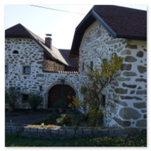 Traditionelles Steinbloßhaus mit rotem Ziegeldach, kleinem Garten, Holzfenstern und Rundbogentor; herbstliche Bäume und gepflasterte Zufahrt am Hi 10 Themenweg