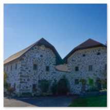 Steinbloßgebäude mit braunem Schindeldach, weißen Fensterrahmen und herbstlicher Gartenumgebung; gepflasterte Zufahrt und Sonnenlicht am Hi 10 Themenweg