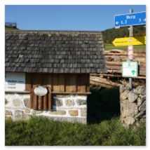 Steinbau mit Infotafel und Rundpfeiler mit Wander- und Radwegschildern; herbstliche Wiese, Holzstapel und klare Beschilderung am Hi 10 Steinbloß-Mauer-Weg bei Berg
