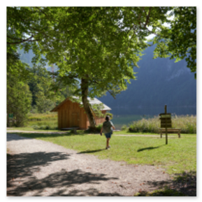 Kleine Holzhütte am See, umgeben von Bäumen und Wiese; Person auf Kiesweg, Holzschild im Gras, Sonnenlicht durch Laub, ruhige Szene vor bewaldetem Hang