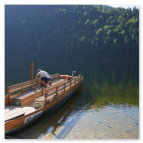 Holzboot mit Person am klaren Seeufer, Steine unter Wasser sichtbar; Waldhang im Hintergrund, Sonnenlicht durch Laub, ruhige Szene mit Natur und Bewegung