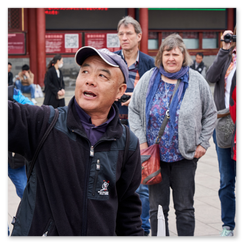 Personengruppe im Freien, zentral ein Mann mit schwarzer Jacke und Baseballkappe, spricht gestikulierend zur Gruppe, umgeben von roten Säulen und chinesischer Architektur, Touristen mit Kameras