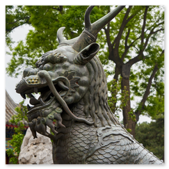 Detailreiche Statue eines mythischen Wesens mit geöffnetem Maul, Hörnern und schuppigem Körper vor traditioneller chinesischer Architektur; im Hintergrund Ziegeldächer, Bäume und ein großer Zierstein, die Szene verbindet symbolische Kraft mit kunstvoller Ruhe.