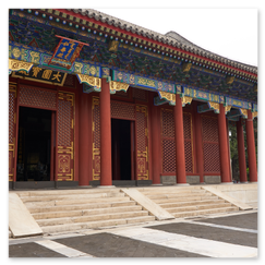 Traditionelles chinesisches Gebäude mit geschwungenem Ziegeldach, roten Säulen und reich verzierten Dachvorsprüngen; vor dem Eingang zwei Tierfiguren auf Sockeln, flankiert von Steinplatten und Grün, die Szene verbindet symbolische Präsenz mit architektonischer Detailfreude.