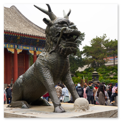Große bronzene Qilin-Figur mit schuppigem Körper, Hörnern und kraftvoller Haltung auf steinernem Sockel; davor eine Kugel, dahinter rotsäuliger Tempel mit goldverzierten Dachvorsprüngen und bemalten Mustern, umgeben von Menschen, die die Szene beleben und die symbolische Präsenz des mythologischen Wesens rahmen.