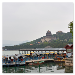 Reihe bunter Tretboote mit gelben, roten und blauen Dächern am Steg des Kunming-Sees; rechts ein Baum mit dichtem Laub und Spaziergänger auf dem Uferweg, im Hintergrund erhebt sich die Halle des Buddhistischen Wohlgeruchs auf dem bewaldeten Hügel, umgeben von traditionellen Gebäuden mit roten Wänden und geschwungenen Dächern, die Szene wirkt ruhig und weit, ein Moment kaiserlicher Gartenkunst.