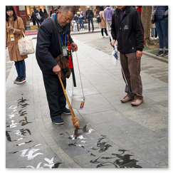 Älterer Mann schreibt mit Wasser und großem Pinsel chinesische Schriftzeichen auf den Steinboden, umgeben von Beobachtern; im Hintergrund rote Säulen, Ziegeldach und traditionelle Architektur, die Szene verbindet vergängliche Kunst mit öffentlicher Aufmerksamkeit und stiller Konzentration.