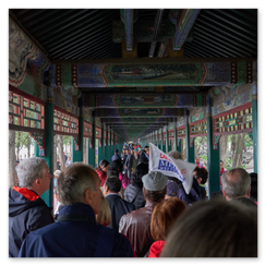 Menschen durchqueren einen reich verzierten Korridor mit bemalten Deckenbalken, roten Säulen und vergitterten Fenstern; draußen Bäume und ein Steinbrückchen über Wasser, die Szene verbindet architektonische Pracht mit lebendiger Bewegung, ein Moment zwischen Kulturdenkmal und Gegenwart.