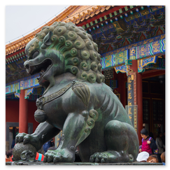 Große Löwenstatue mit gelockter Mähne und Pranken auf Kugel vor traditionellem chinesischem Gebäude mit roten Säulen und farbig verzierten Dachvorsprüngen; umgeben von Besuchern, die Szene verbindet symbolische Stärke mit kaiserlicher Architektur und lebendiger Gegenwart.