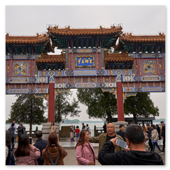 Dreiteiliger chinesischer Paifang mit roten Säulen, mehrstöckigen Dächern und farbig verzierten Ornamenten; mittig ein Schriftfeld mit gelben Zeichen auf blauem Grund, davor Menschen mit Kameras, im Hintergrund Bäume und Wasser, die Szene verbindet festliche Architektur mit öffentlicher Lebendigkeit.