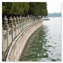 Reihe kunstvoll verzierter Steinpfosten mit dekorativen Abschlüssen entlang eines Uferwegs; dahinter ruhiges, grünlich schimmerndes Wasser mit einem überdachten Boot in der Ferne, hohe Bäume spenden Schatten, die Szene wirkt still und ausgewogen, ein Moment zwischen Handwerk und Natur.
