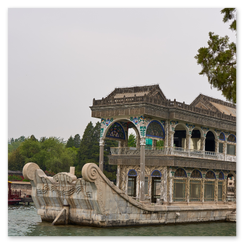 Prunkvoll gestaltetes Marmorboot mit geschwungenem Dach und farbigen Ornamenten am Rand des Kunming-Sees; dahinter traditionelle Gebäude mit gelben Ziegeldächern und roten Wänden, umgeben von Bäumen und ruhigem Wasser, die Szene verbindet kaiserliche Gartenarchitektur mit stiller Eleganz und historischer Symbolik.