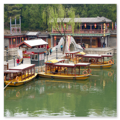 Gelbdachige Holzboote am ruhigen Kanal neben einem Steg; gegenüber traditionelle Häuser mit roten Laternen und grauen Ziegeldächern, eine kleine Steinbrücke verbindet die Ufer, die Szene wirkt still und lebendig, ein Moment chinesischer Wasserstadtkultur.