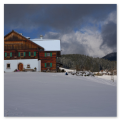 Schneebedecktes Holzhaus am Hang, Kirche von Gosau im Hintergrund, bewölkter Himmel