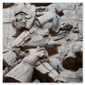 Zerbrochene Tonfiguren der Terra-Cotta-Armee in Xi’an, liegend auf steinernem Boden. Einzelteile wie Köpfe, Arme und Torsos sind verstreut; die Figuren tragen Rüstung oder Gewänder. Die Szene zeigt den Zustand vor oder während der Restaurierung.