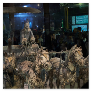 Ausstellungsstück im Museum in Xi’an: Lebensgroßes Modell eines antiken Streitwagens mit vier Tonpferden und einer Figur in traditioneller Kleidung. Das Ensemble steht in einer Glasvitrine; Besucher betrachten die Szene im Hintergrund. Beleuchtete Infotafeln ergänzen die Präsentation.