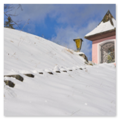Verschneiter Hang mit Kapelle und Laterne, Aufstiegspfad im Schnee, Nadelbäume im Hintergrund