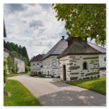 Mehrere Steinbloßhäuser mit dunklen Horizontalstreifen und Ziegeldächern entlang geschwungener Straße; Baumkrone, gepflegte Grünflächen und Waldhintergrund in Katzing bei Rohrbach-Berg im Mühlviertel