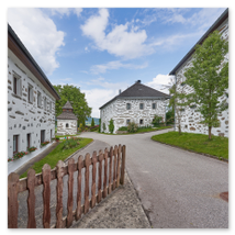 Traditionelle Steinbloßhäuser mit dunkler Musterung, Ziegeldächern und Blumenschmuck entlang gepflasterter Straße; Lattenzaun, Grünflächen und Bäume in Katzing bei Rohrbach-Berg im Mühlviertel