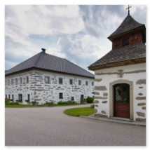 Steinbloßhaus mit dunklem Ziegeldach neben kleiner Kapelle mit Kreuz und Glockenturm; gepflegte Grünflächen, gepflasterte Straße und ruhige Dorfszene in Katzing bei Rohrbach-Berg im Mühlviertel