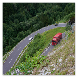 Drei Fahrzeuge – ein roter Sattelschlepper, ein silberner PKW und ein dunkles Auto – fahren auf einer kurvigen Bergstraße durch dicht bewaldetes Gebiet. Die Straße verläuft in einem U-förmigen Bogen und ist mit Leitplanken und gelben Richtungsschildern gesichert. Im Vordergrund liegt ein steiniger Hang mit grüner Vegetation. Die Aufnahme stammt von einem erhöhten Standpunkt und zeigt die Szene bei diffusem Tageslicht.
