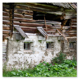 Oberer Gebäudeteil aus waagrecht gestapelten Holzstämmen, teilweise beschädigt mit einer großen Öffnung. Dach mit Holzschindeln, teils fehlend oder verwittert. Unterer Gebäudeteil aus Stein oder Beton mit drei kleinen vergitterten Fenstern und geöffneten Holzläden. Vor dem Gebäude grüne Vegetation und Gras.