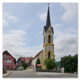 Gelb-weiße Kirche mit hohem Turm und Uhr im Zentrum einer kleinen Ortschaft. Umgeben von Wohn- und Geschäftsgebäuden, darunter ein rotes Haus mit Beschriftung Schwarzwald Tourismus und Haus des Gastes. Die Straße verläuft bogenförmig um die Kirche. Im Hintergrund grüne Hügel und Bäume, teils bewölkter Himmel.