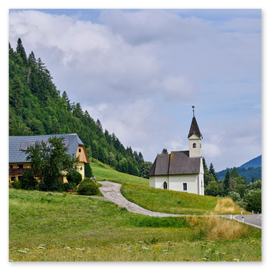 Weiße Kirche mit hohem Turm und Kreuz an der Spitze in einem kleinen Dorf, gelegen in einem grünen Tal. Neben der Kirche mehrere traditionelle Gebäude mit Satteldächern. Eine kurvige Straße führt durch den Ort. Umgeben von bewaldeten Hügeln, teils bewölkter Himmel, weiches Tageslicht.