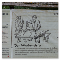 Informationsschild beim Erlebnis Burgbau Friesach zeigt eine Illustration von zwei Handwerkern in mittelalterlicher Kleidung, die an einem Holzrahmen arbeiten. Der Text erklärt die Rolle des Werkmeisters im 14. Jahrhundert als organisatorische Leitung der Baustelle.