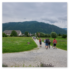 Menschen in Freizeitkleidung gehen auf einem Schotterweg durch eine Wiese, im Hintergrund Fahnen und Häuser. Die Szene zeigt den Moment nach dem Besuch des mittelalterlichen Burgbauprojekts in Friesach, eingebettet in eine ländliche Umgebung mit bewaldeten Hügeln