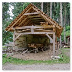Ein rustikaler Holzschuppen mit Schindeldach steht am Rand eines Waldes beim mittelalterlichen Burgbau in Friesach. Im Inneren sind Holzplanken ordentlich gestapelt, der Boden ist naturbelassen und ein Pfad führt zum Eingang. Die Szene zeigt traditionelle Bauweise und vermittelt die Atmosphäre eines historischen Handwerksplatzes, an dem ohne moderne Maschinen gearbeitet wird.