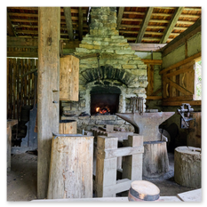 Das Bild zeigt die Innenansicht einer traditionellen Schmiede beim Burgbauprojekt in Friesach. Im Zentrum steht ein offener Steinofen mit brennendem Feuer, umgeben von Amboss, Hämmern und weiteren Werkzeugen des Schmiedehandwerks. Die Werkstatt ist aus Holz gebaut, mit rustikalen Balken und einem offenen Bereich, durch den Tageslicht einfällt. Die Szene vermittelt eindrucksvoll, wie mit mittelalterlichen Methoden und ohne moderne Technik gearbeitet wird.