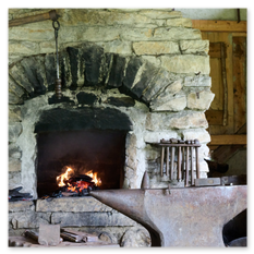 Ein brennender Steinofen steht im Zentrum einer historischen Schmiedewerkstatt beim Burgbauprojekt in Friesach. Über dem Feuer hängt ein Metallstab, umgeben von Zangen und Werkzeugen an der Steinwand. Vor dem Ofen liegt ein massiver Amboss, bereit für die Bearbeitung glühender Metalle. Die Werkstatt mit Holzträgern und rustikalen Mauern vermittelt die Atmosphäre mittelalterlicher Handwerkskunst, wie sie seit Jahrhunderten praktiziert wurde.